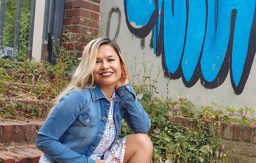 Eine Frau sitzt auf einer Treppe. Sie sieht freundlich aus und trägt eine Jeansjacke. Im Hintergrund sieht man ein blaues Graffiti.