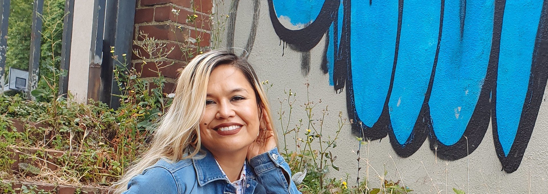 Eine Frau sitzt auf einer Treppe. Sie sieht freundlich aus und trägt eine Jeansjacke. Im Hintergrund sieht man ein blaues Graffiti.