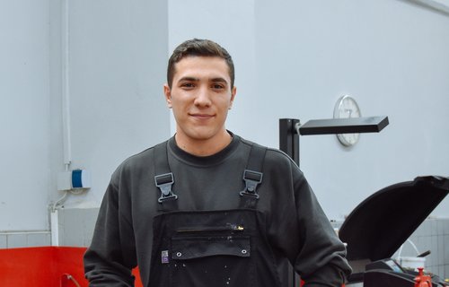 Ein junger man steht in einer Werkstatt. Er trägt Arbeitskleidung und sieht glücklich aus.