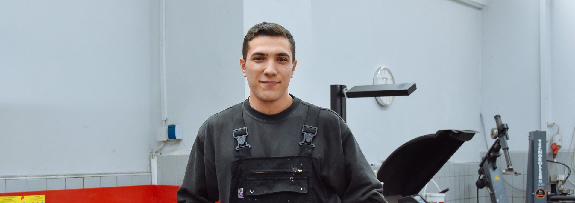 Ein junger man steht in einer Werkstatt. Er trägt Arbeitskleidung und sieht glücklich aus.