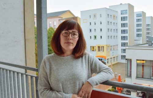 Eine junge Frau steht auf ihrem Balkon. Sie schaut ernst und es ist schlechtes Wetter.