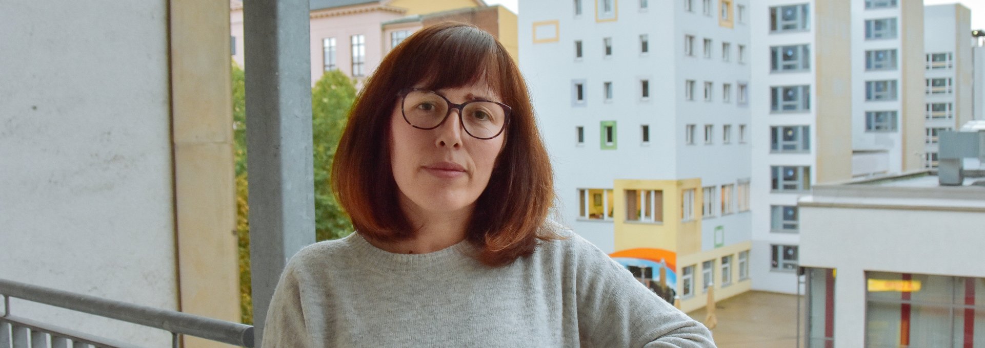 Eine junge Frau steht auf ihrem Balkon. Sie schaut ernst und es ist schlechtes Wetter.