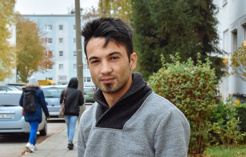 Ein sehr junger Mann aus Syrien steht vor einem Wohnblock. Er schaut ernst.