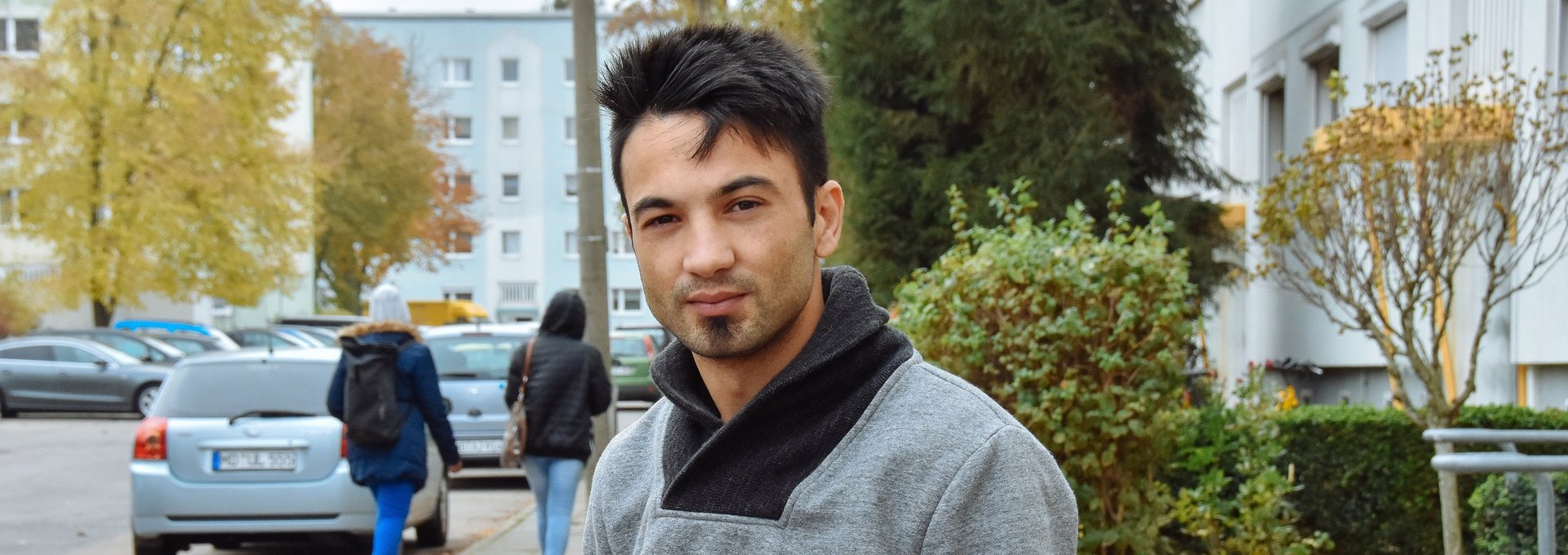 Ein sehr junger Mann aus Syrien steht vor einem Wohnblock. Er schaut ernst.