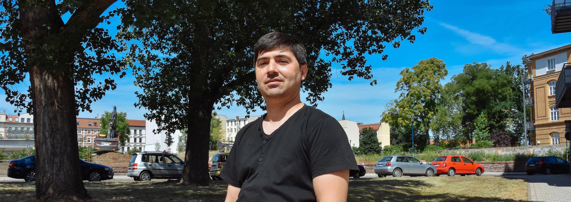 Ein freundlicher Mann steht an einen Stein gelehnt. Er trägt ein schwarzes T-Shirt und eine blaue Jeans.