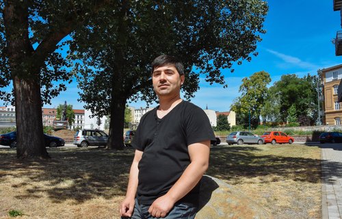 Ein freundlicher Mann steht an einen Stein gelehnt. Er trägt ein schwarzes T-Shirt und eine blaue Jeans.