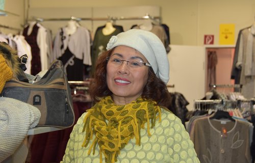 Eine ältere Dame steht in ihrem Modegeschäft und trägt einen großen Hut.