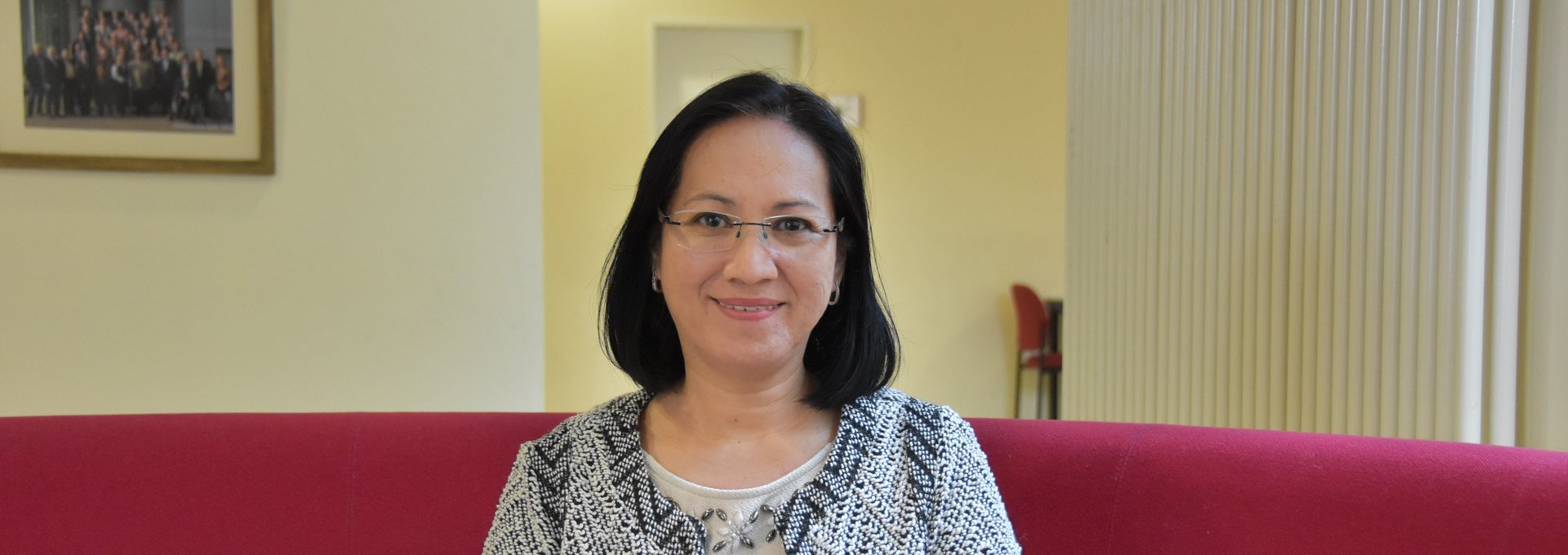 Eine asiatische Frau sitzt auf einem Sofa. Sie trägt eine Brille und eine karierte Weste.
