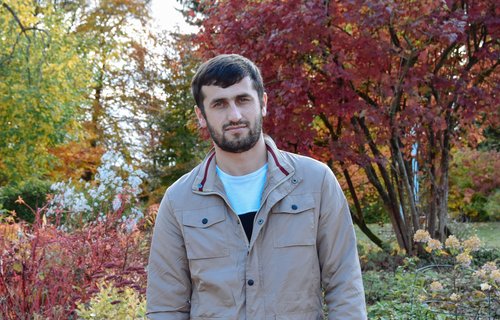 Ein junger Mann mit Vollbart steht in einem Park. Die Bäume haben bunte Blätter.