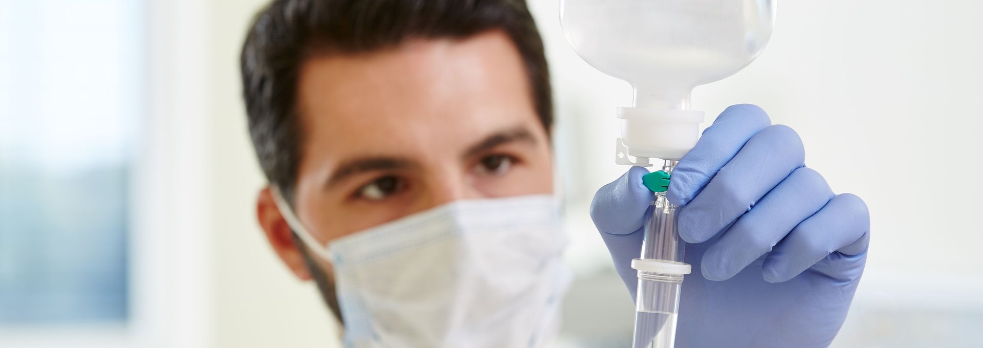 Das Foto zeigt einen jungen Mann bei einer pflegerischen Tätigkeit im Krankenhaus. Mit Mundschutz und Handschuhen stellt der die Fließrate an einem Tropf ein.