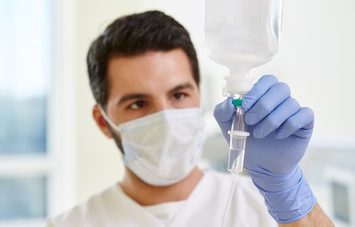 Das Foto zeigt einen jungen Mann bei einer pflegerischen Tätigkeit im Krankenhaus. Mit Mundschutz und Handschuhen stellt der die Fließrate an einem Tropf ein.