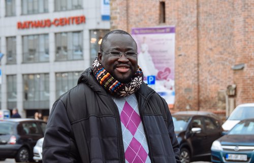 Ein junger schwarzer Mann steht auf einem Markplatz. Er trägt dicke Wintersachen.