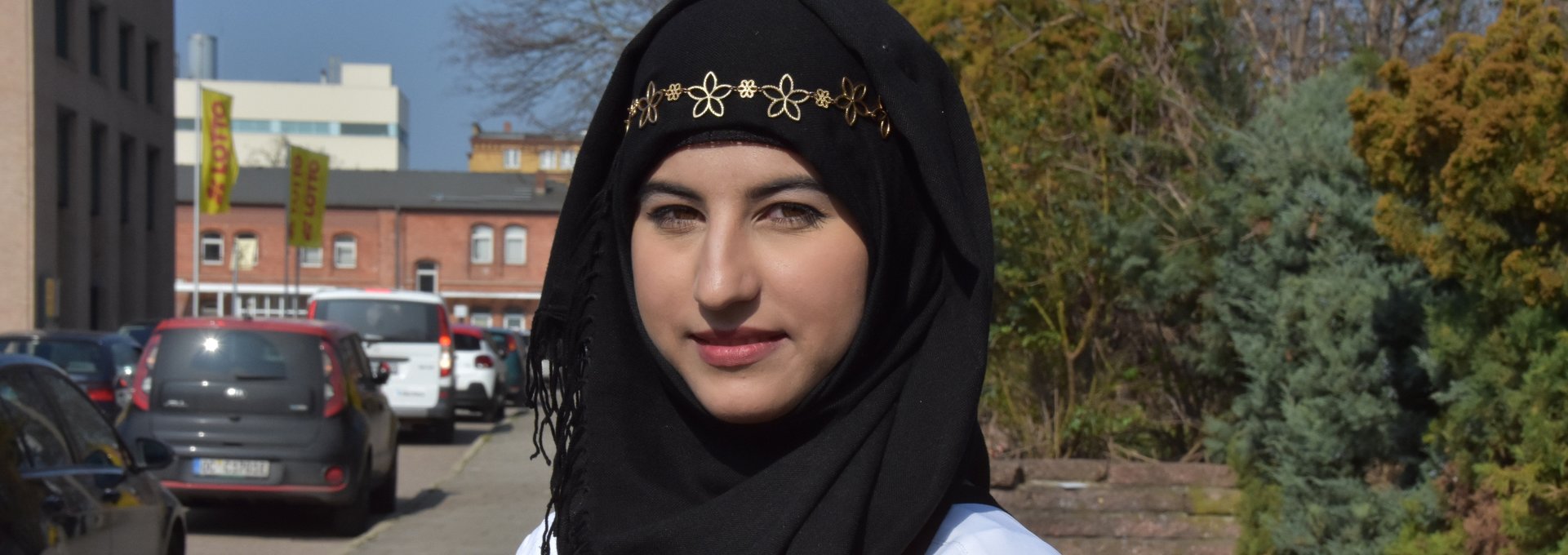 Ein junges Mädchen steht auf der Straße. Sie trägt ein schwarzes Kopftuch mit goldenem Schmuck.