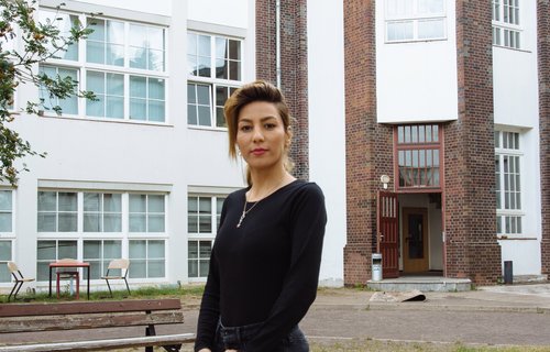 Eine junge Frau steht in einem Innenhof. Sie ist schwarz gekleidet und stark geschminkt.