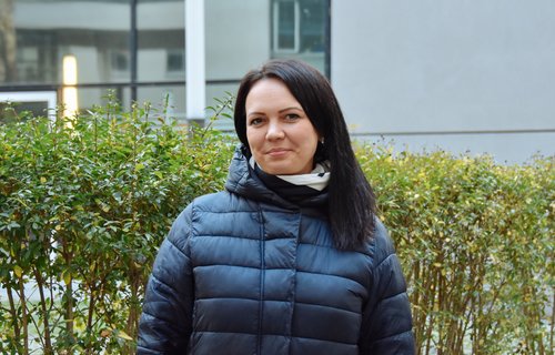 Ein junge Frau steht in Winterjacke vor einem Busch. Sie hat schwarze Haare und lächelt.