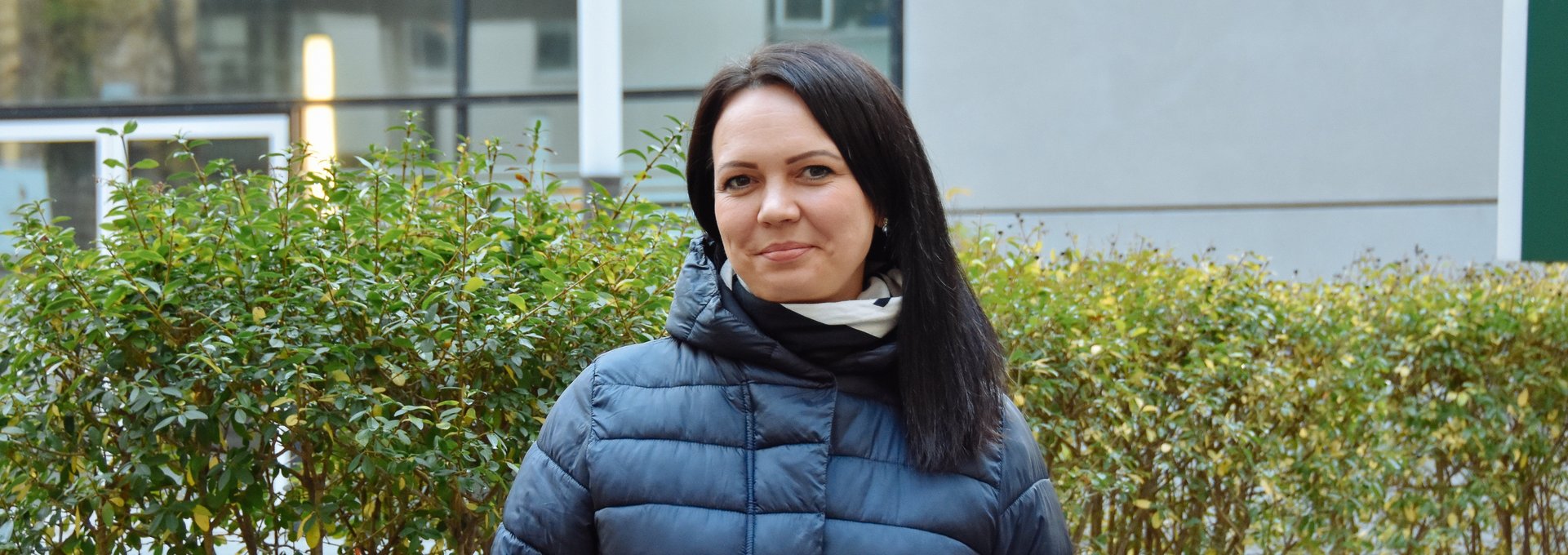 Ein junge Frau steht in Winterjacke vor einem Busch. Sie hat schwarze Haare und lächelt.