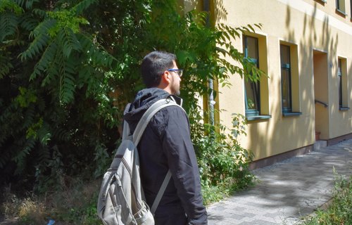 Ein junger Mann steht mit dem Rücken zur Kamera. Er hat einen Rucksack auf dem Rücken und trägt eine Brille.