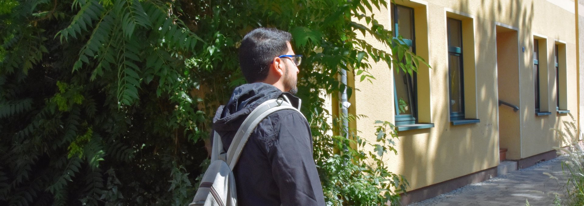Ein junger Mann steht mit dem Rücken zur Kamera. Er hat einen Rucksack auf dem Rücken und trägt eine Brille.
