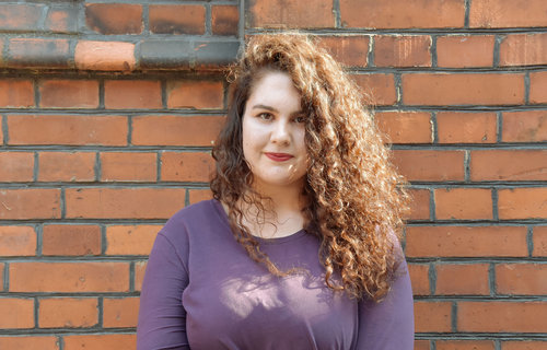 Eine junge Frau steht vor einer roten Steinwand. Sie hat langes , braunes Haar mit Locken.
