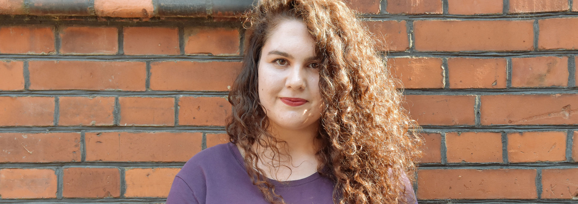 Eine junge Frau steht vor einer roten Steinwand. Sie hat langes , braunes Haar mit Locken.
