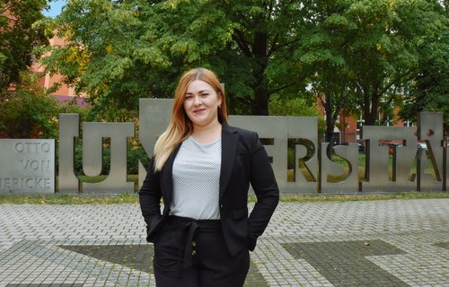 Eine junge Frau steht auf einem Platz. Sie hat blonde Haare und lächelt.
