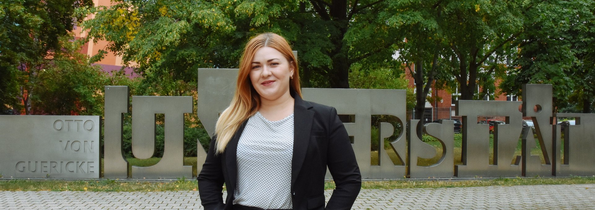 Eine junge Frau steht auf einem Platz. Sie hat blonde Haare und lächelt.